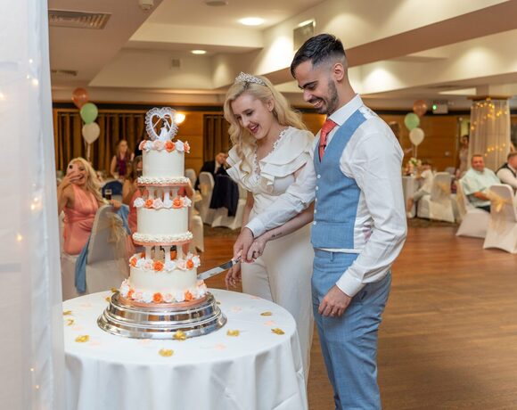 Bride in white gown and groom in blue suit cut a three-tier cake with peach roses in a hotel ballroom