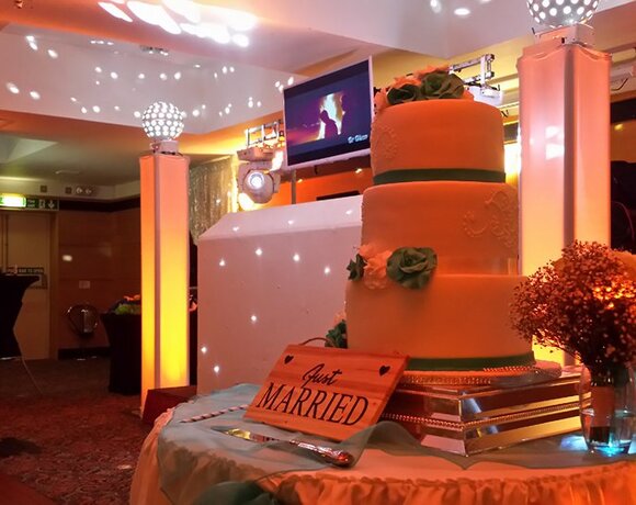Three-tier wedding cake with green sugar roses and 'Just Married' sign on a lit reception table in warm orange event room.