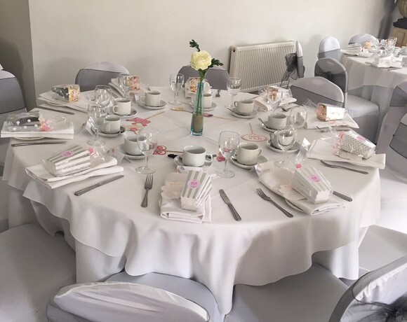 Round table set for a baby shower with white linens, teacups, favors, gray organza chair bows and single white rose.