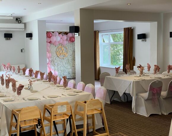 Bright dining room set for a baby shower with white tables, pink napkins, chair sashes and floral 'Oh Baby' balloon wall.