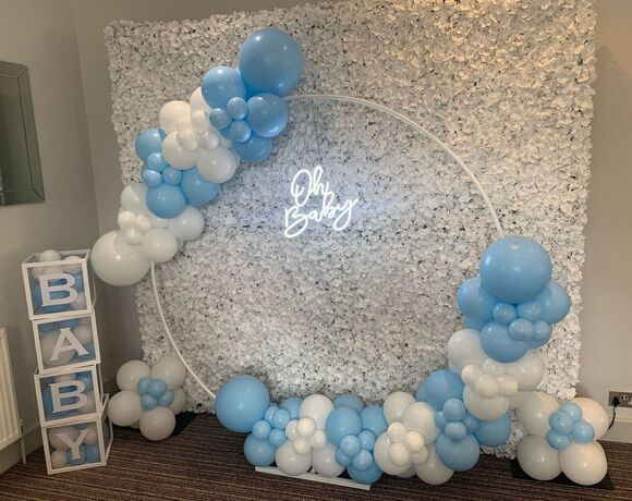 White floral backdrop with neon 'Oh Baby' sign, pale blue-and-white balloon arch and stacked BABY blocks for baby shower.