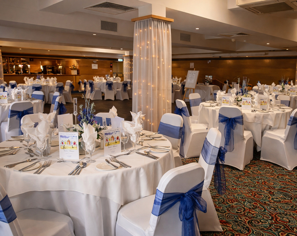 Elegant banquet hall set for wedding reception: round tables with white linens, navy-blue chair sashes, floral centerpieces and fairy-lit columns.