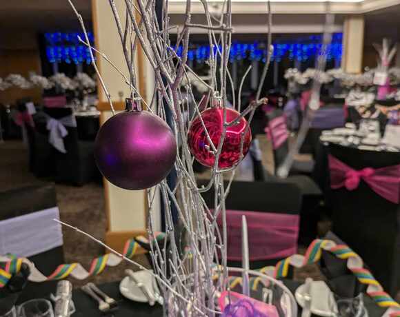 Silver-branch centerpiece with purple and magenta baubles over a dressed banquet table — black chairs with pink sashes, glassware and blue lights.