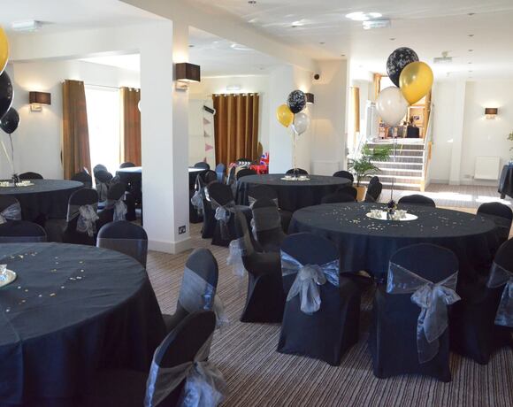 Banquet room set for celebration with round tables draped in black linens, silver-sashed chairs and gold/black balloons.