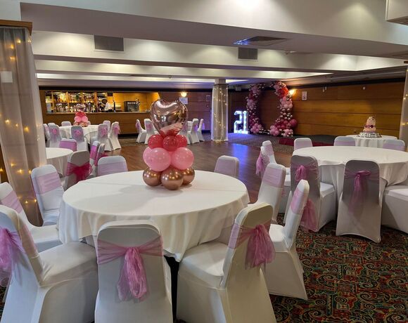 Banquet room set for a 21st: round tables with white linens, chairs tied with pink bows, heart balloon centerpieces, balloon arch, lit '21' and cake
