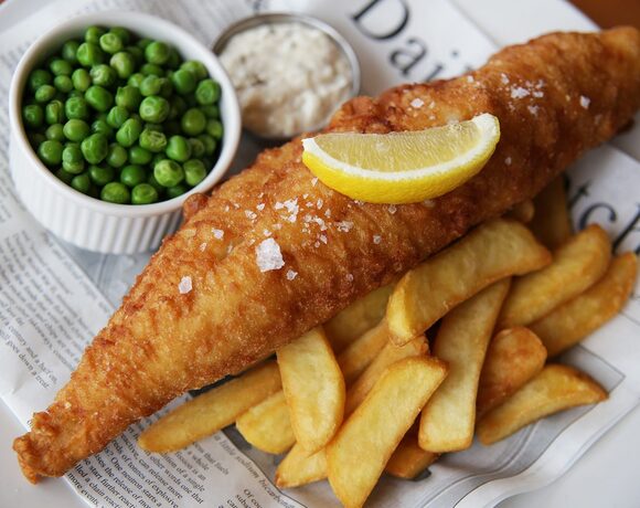Crispy beer-battered fish with sea salt and lemon wedge, thick-cut chips and a bowl of peas with tartar sauce on newspaper-lined plate.