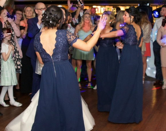 Bridesmaids in navy lace gowns dancing at lively hotel wedding reception, guests clapping around a wood dance floor.