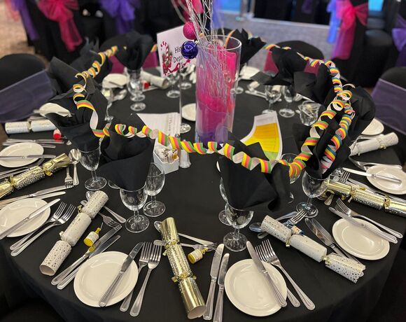 Banquet table set for a festive dinner with black linens, black napkins, place settings, party crackers, colorful streamers and pink centerpiece.