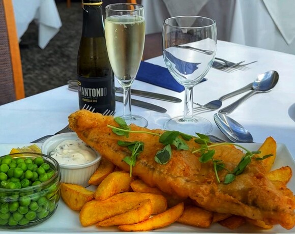 Crispy beer-battered fish with chunky chips, garden peas and tartare, paired with a chilled Prosecco flute on a white table.