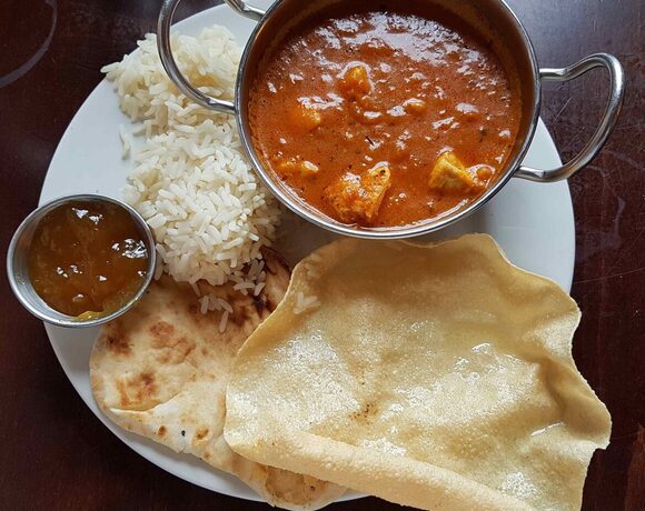 Creamy tomato chicken curry in a metal bowl with basmati rice, warm naan, crisp papadum and mango chutney
