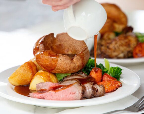 Sliced roast beef with Yorkshire pudding, golden roast potatoes, glazed carrots and greens as gravy is poured