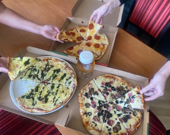 Three large pizzas in open boxes, pepperoni, mushroom with olives, and pesto cheese, hands grabbing slices around frothy beer.