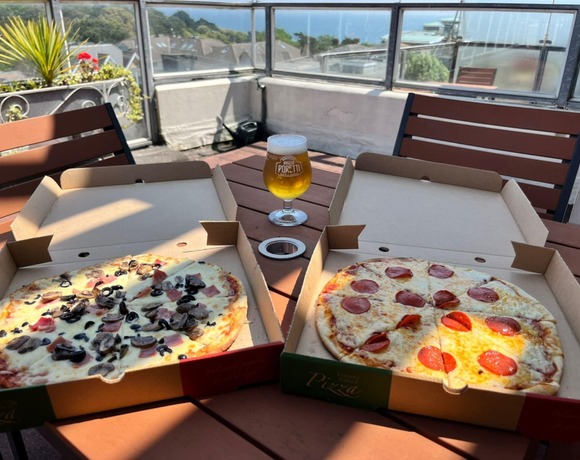 Rooftop terrace dining with two open pizza boxes (pepperoni and mushroom/olive), a beer glass on wooden table and sea view.
