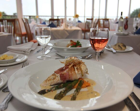 Fine-dining hotel restaurant with bacon-wrapped chicken, asparagus in cream sauce, rosé glass, white-linen table and bright terrace windows.