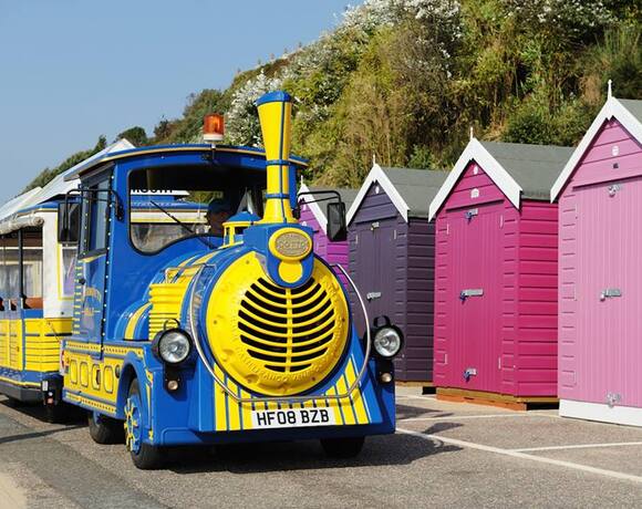 Bright blue-and-yellow tourist train beside pink and purple beach huts on a sunny seaside promenade for family rides