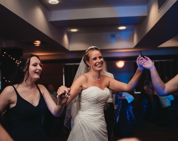Bride in strapless gown and veil dancing with bridesmaids on a hotel ballroom floor, warm lighting and joyful reception atmosphere