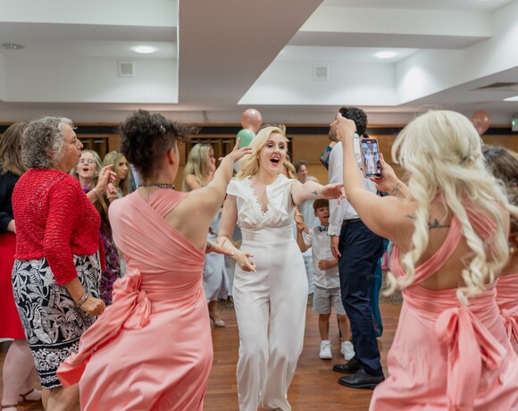 Bride in white jumpsuit dancing on a hotel reception dance floor, bridesmaids in pink, guests filming and balloons
