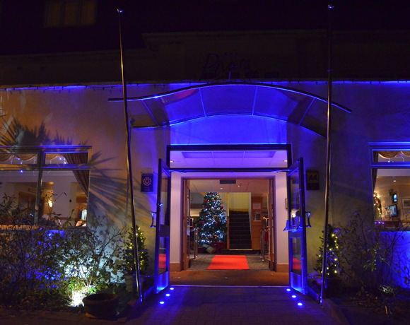 Hotel entrance with glass canopy and blue uplighting, open doors to a red carpet and decorated Christmas tree in the lobby.