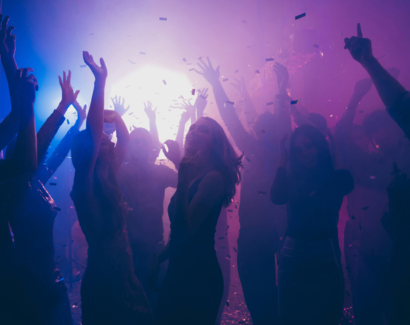 Lively hotel nightclub dance floor with guests dancing under purple and blue lights, confetti falling and raised hands.