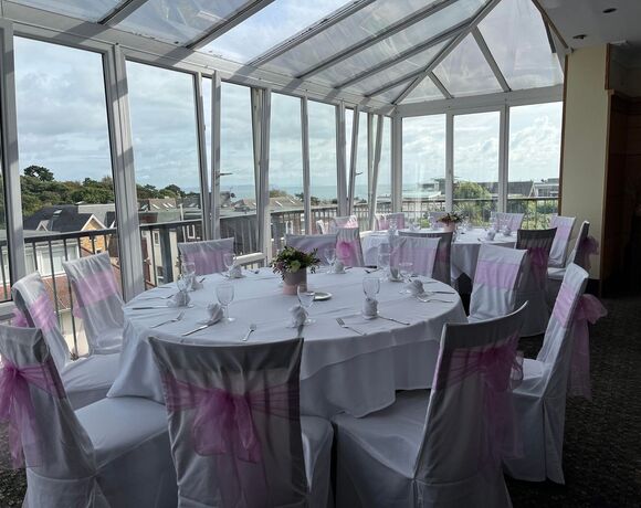 Bright conservatory dining room with round tables in white linens, pink-sashed chairs, floral centerpieces and sea view.