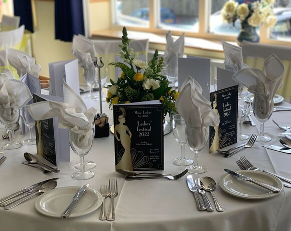 Elegant table with white linens, napkins folded in glasses, yellow-white floral centerpiece and Ladies' Festival 2022 menus.