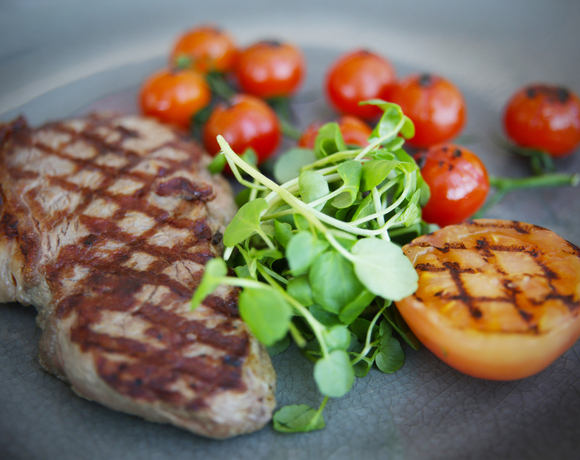 Grilled steak with cross-hatch sear, charred cherry tomatoes, grilled tomato half and fresh microgreens on a slate plate.