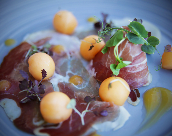 Thin prosciutto carpaccio and melon balls with pea shoots and microgreens on a blue plate, drizzled citrus sauce
