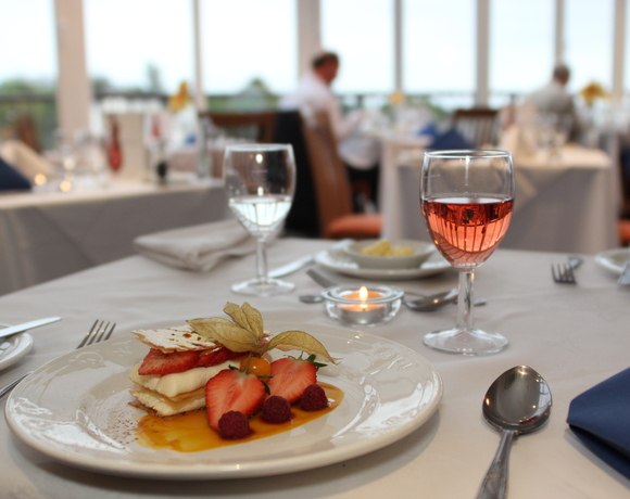 Elegant restaurant table with layered strawberry dessert, glass of rosé, water glass and lit candle on a white tablecloth by large windows.