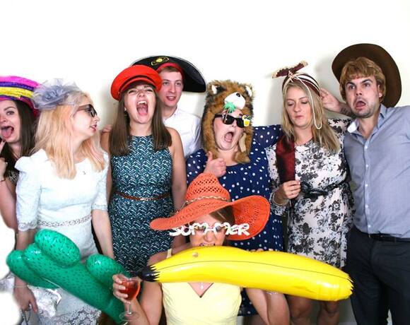 Eight people in formal attire wearing playful hats and holding inflatable banana and cactus, laughing and posing in a photo booth.