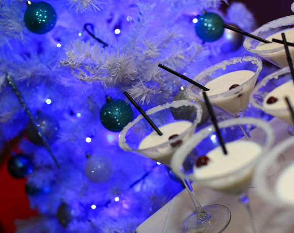 Creamy holiday cocktails in sugar-rimmed martini glasses with cherries and black straws next to white Christmas tree lit blue.