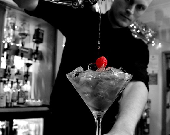 Bartender pouring a shaken cocktail from a steel shaker into a chilled martini glass with ice and a fresh raspberry garnish at hotel bar.