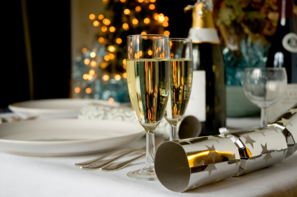 Festive hotel dining table with champagne flutes, bottle and crackers on white linen, blurred holiday tree lights behind