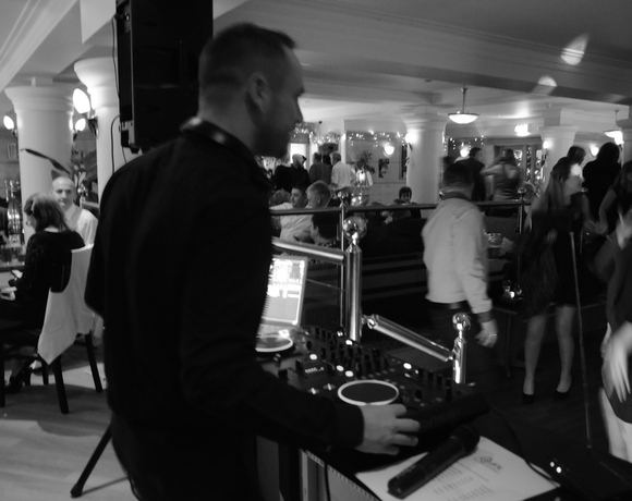 Hotel lounge holiday party with DJ at console, guests dining and dancing among columns, warm lamps and a decorated Christmas tree.