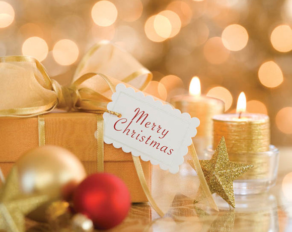 Festive holiday gift with 'Merry Christmas' tag, gold ribbon, ornaments and lit candles creating warm bokeh lobby ambiance.
