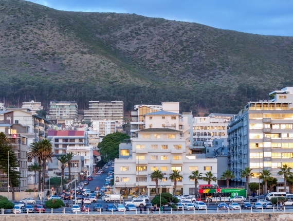 Riviera Suite with balconies and palm trees, lit windows, parked cars on a busy coastal promenade with mountain backdrop at dusk.