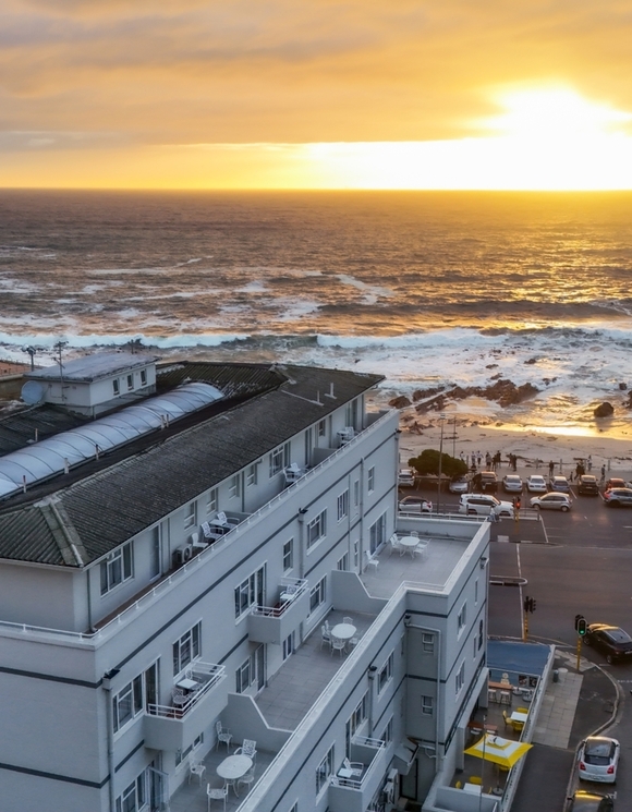 Riviera Suite with private balcony tables and terraces overlooking a rocky beach and golden sunset over the sea.
