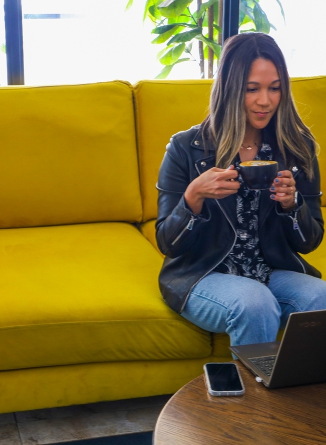 Guest on bright yellow lounge sofa sipping coffee while using laptop at a cozy, plant-filled hotel lobby workspace.