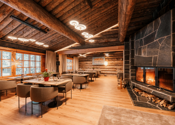 Cozy rustic dining room with wooden log walls, modern chandeliers, large windows with snow views, and a stone fireplace with stacked firewood