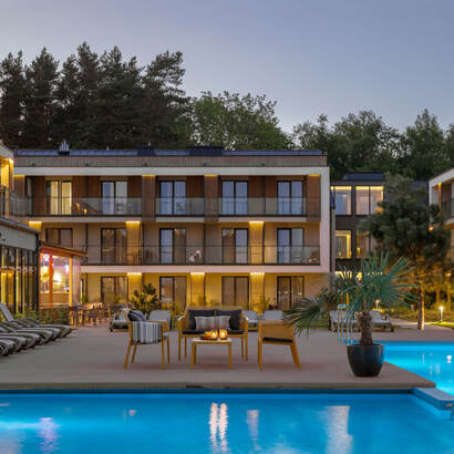 Außenpool bei Dämmerung mit Liegen, Lounge-Sitzgruppen und beleuchteten, modernen Hotelbalkonen vor einer Baumkulisse