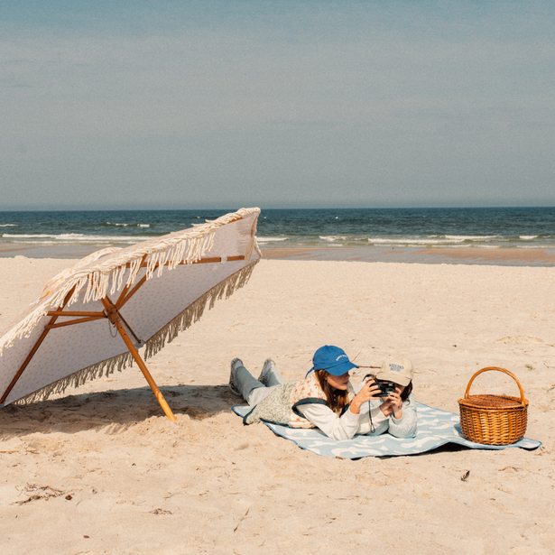 Couple relaxing under fringed beach umbrella on a towel, taking photos beside a picnic basket on a quiet sandy shore