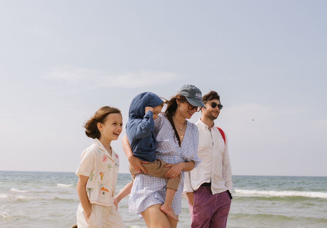Rodzina idąca boso po słonecznej piaszczystej plaży; mama niesie malucha, uśmiechnięte dzieci i pies przy brzegu