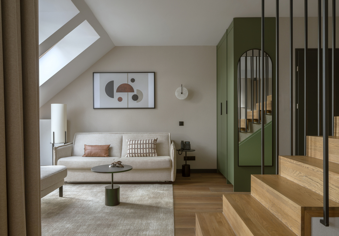 Przytulny salon w apartamencie ze świetlikiem, beżową sofą i poduszkami, abstrakcyjną grafiką, lampą stojącą, zieloną szafą z lustrem i drewnianymi schodami.