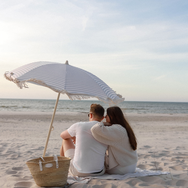 Couple cuddling on a blanket under a striped beach umbrella, straw tote beside them, calm sea at sunset