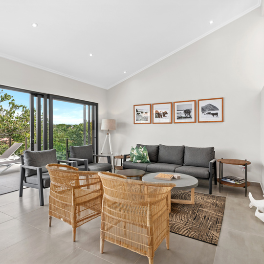 Bright living area with modern gray sofa, wicker chairs, coffee tables, and open sliding doors to a balcony with lounge chairs and lush greenery views
