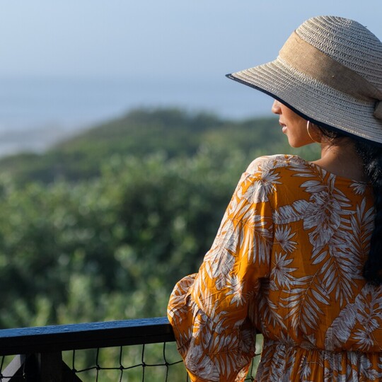 Guest in a floral dress and sun hat enjoying serene ocean and lush green hillside view from hotel balcony