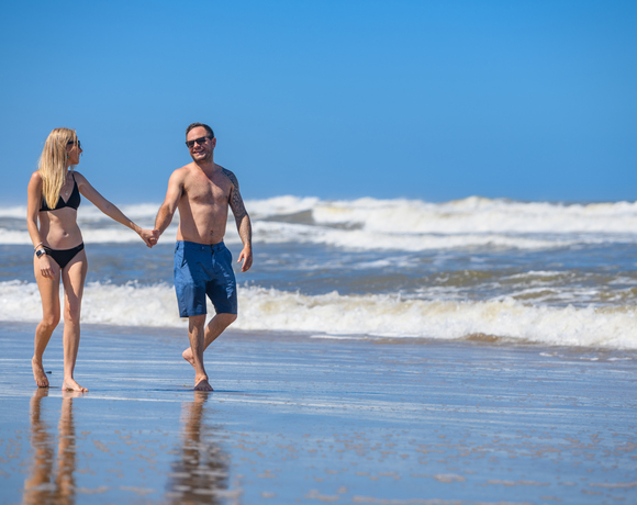 Couple holding hands and walking barefoot along the beach with waves and clear blue sky on a sunny day
