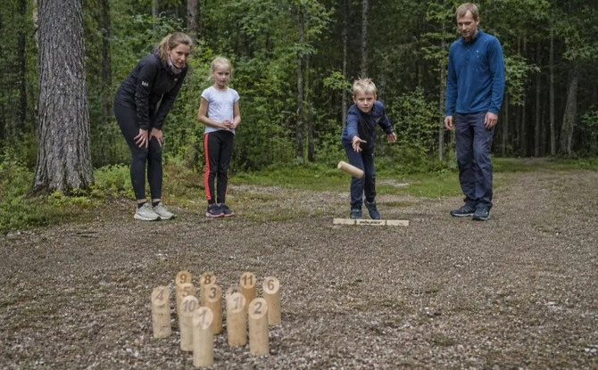 Family playing a wooden throwing lawn game in a forest clearing, parents and children enjoying outdoor activities.