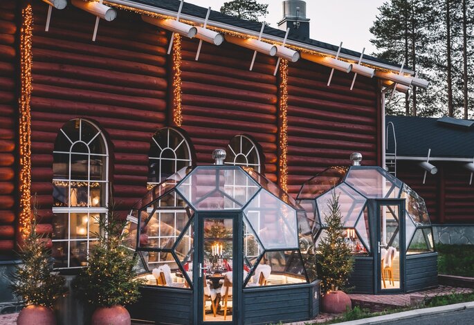 Yksityisillallisiin tarkoitetut, jouluvaloin valaistut lasi-iglut perinteisessä punaisessa hirsirakennuksessa toimivan Three Elves -ravintolan vieressä Santa Claus Holiday Villagessa Rovaniemellä