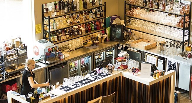 Hotel bar with bartender preparing cocktails, stocked spirits, glass racks and chilled fridges behind a wooden island counter.