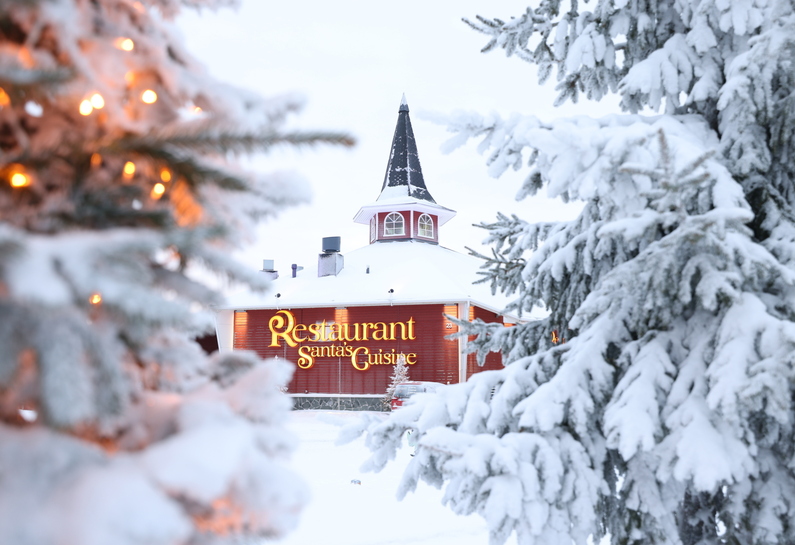 Santa’s Cuisine -ravintolan valaistu kyltti ja torni lumisten kuusten kehystäminä Santa Claus Holiday Villagessa Rovaniemellä